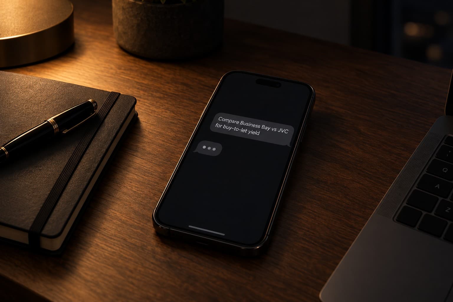 A phone on a desk at night with a broker's question on screen: Compare Business Bay vs JVC for buy-to-let yield.