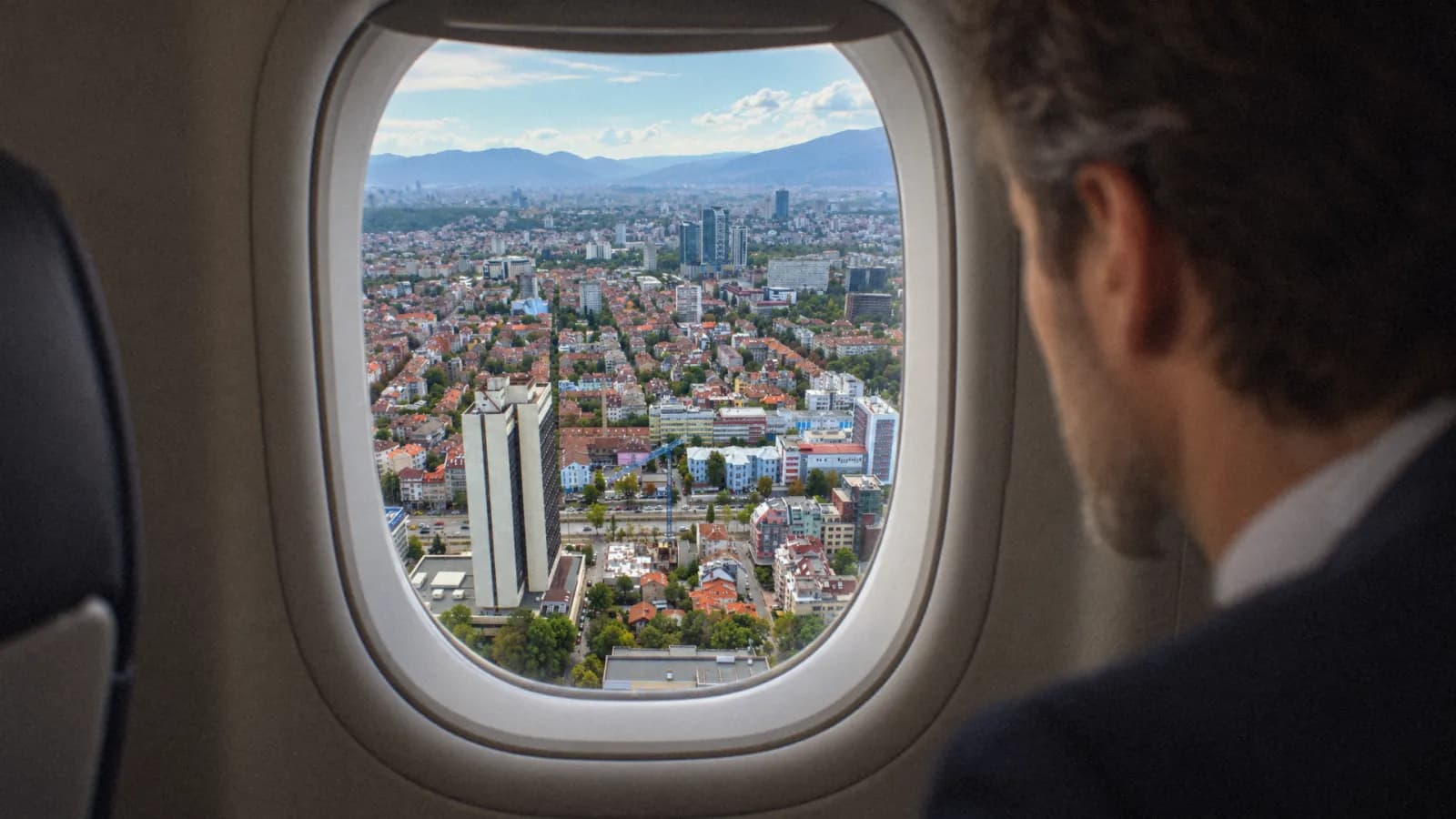 A man in a dark suit looking out of an aircraft window at the city below, the tower visible among the blocks.