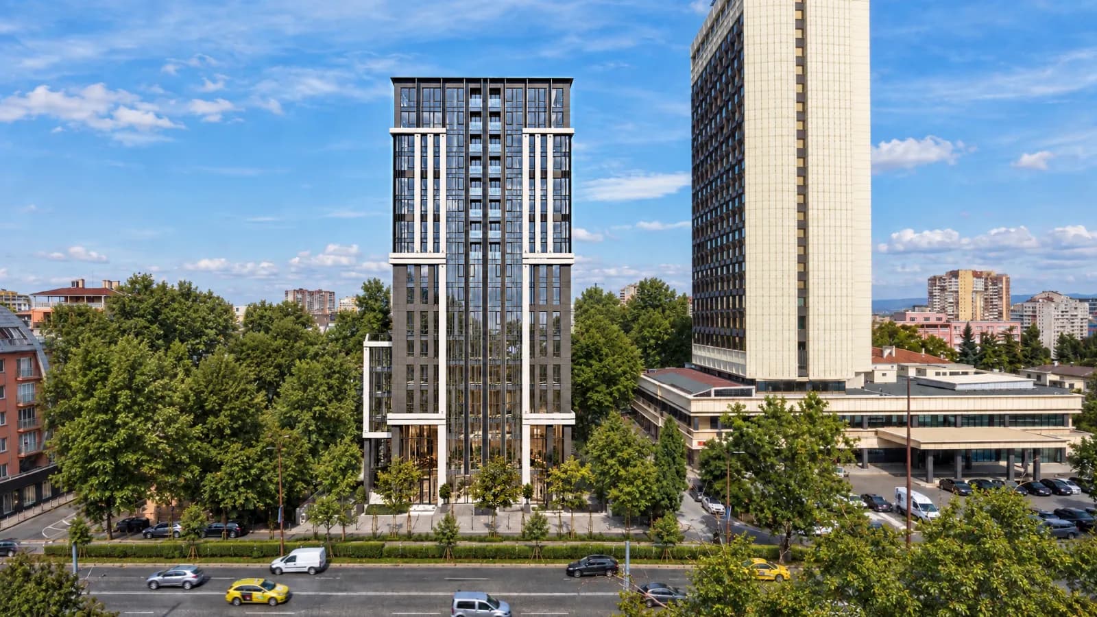 The tower in full daylight from across a busy avenue, a dark vertical facade between trees, a taller white hotel block to its right and low city blocks behind.