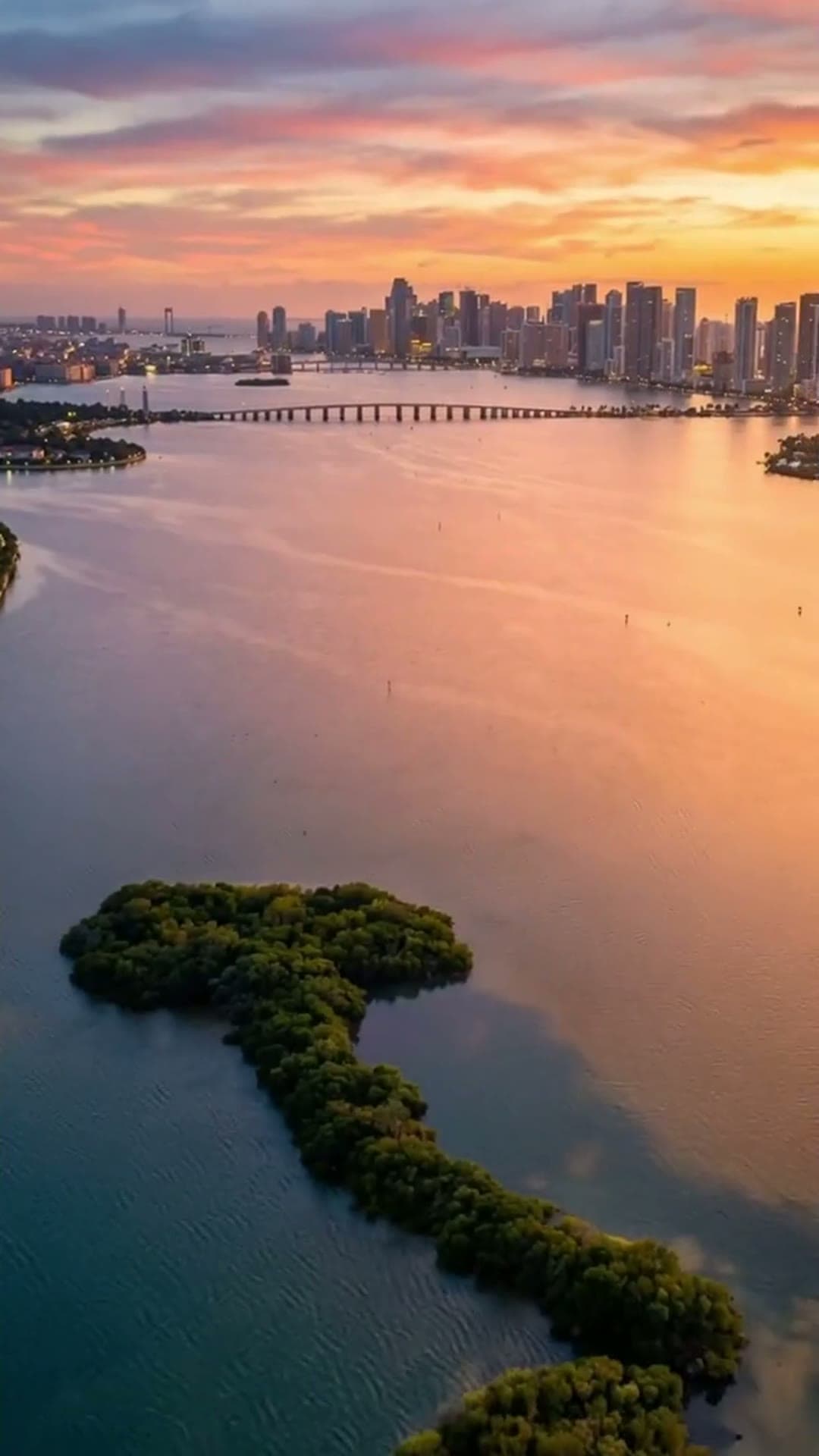 An AI-generated aerial flythrough over a bayfront skyline at golden-hour sunset, mangrove islands threading the foreground water, warm orange light across the towers.
