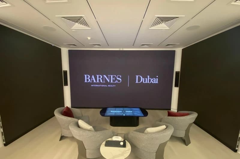 The BARNES Dubai immersive room as it stands today: a wall-width screen showing the BARNES International Realty and Dubai wordmarks, a touchscreen table in front of it running the same lockup, and four grey armchairs with red cushions around a small marble side table.