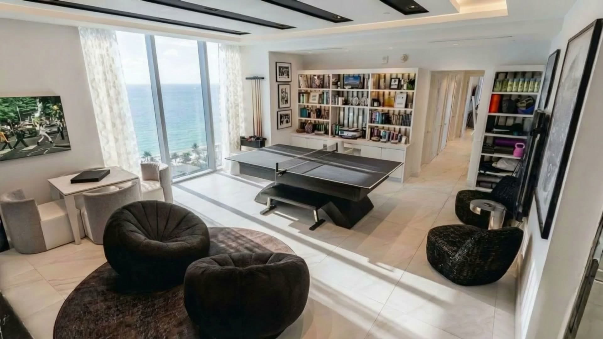 A beachfront game room with white shelving full of books, a black table-tennis table and two round black chairs, the sea through floor-to-ceiling windows.