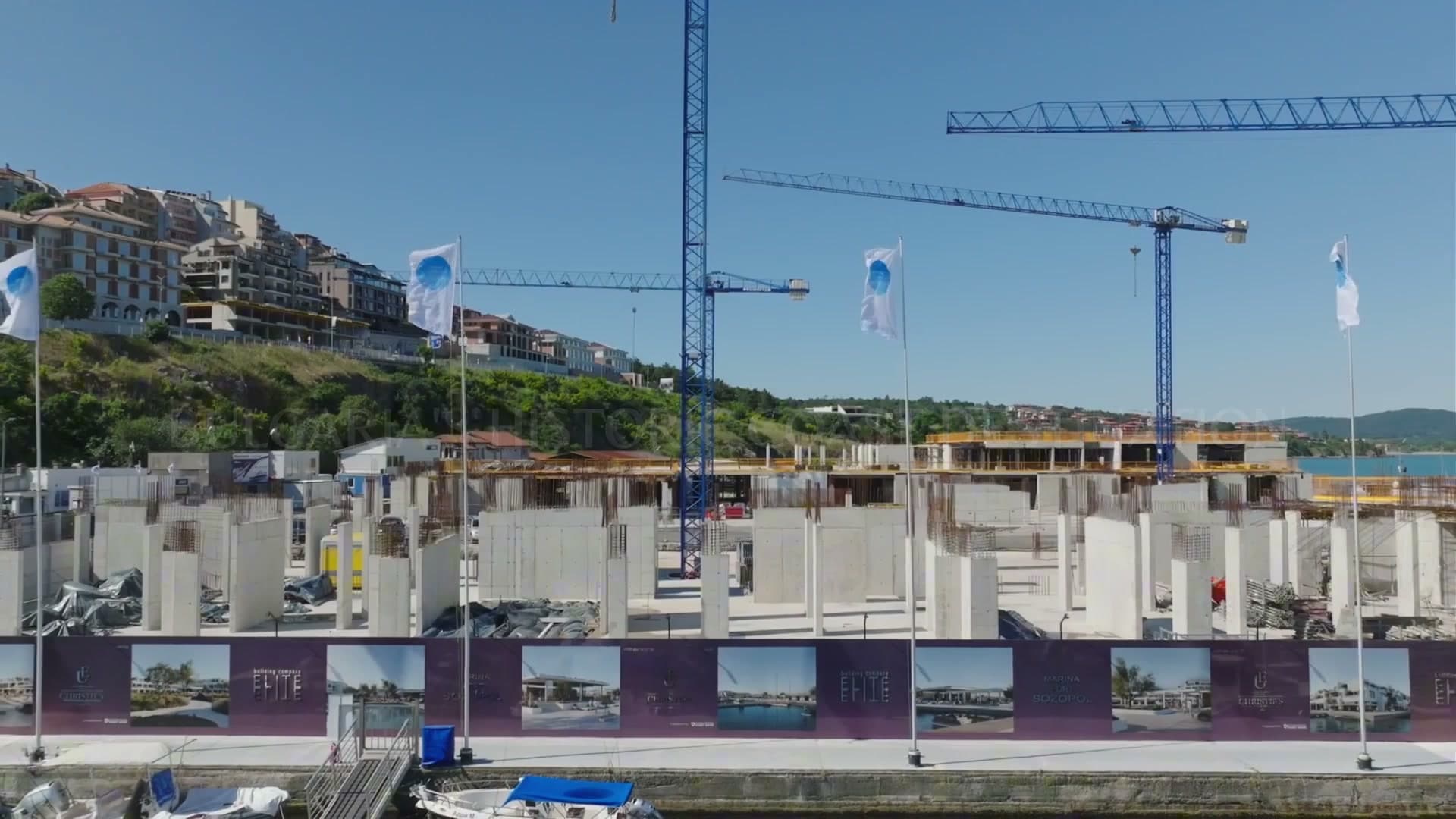 The construction site at street level: tower cranes, raw concrete walls and a printed hoarding along the quay.