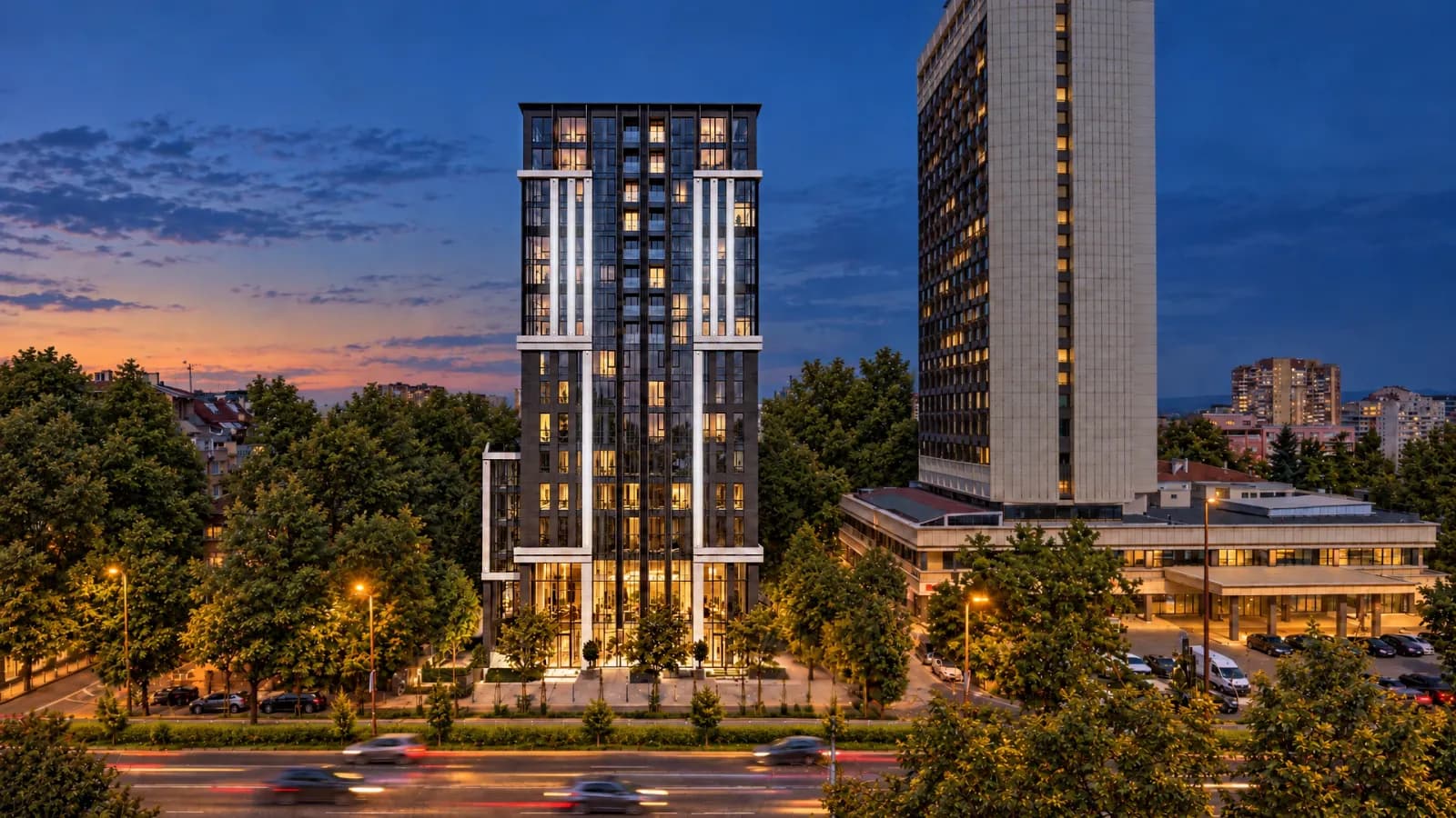A slim residential tower with a dark vertical facade at dusk, its windows lit warm against a deep blue sky, trees and a lit avenue in front and a taller white hotel block to the right.