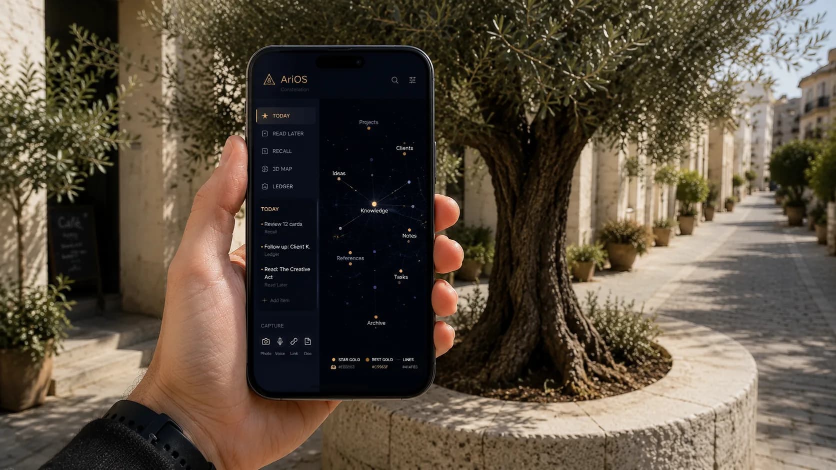 A hand holding a phone on a sunlit Mediterranean street lined with olive trees, the screen showing AriOS's Constellation web app: a sidebar listing Today, Read Later, Recall, 3D Map, and Ledger, a Today list with sample tasks, a Capture row with Photo, Voice, Link, and Doc options, and a 3D knowledge-map of connected nodes (Knowledge, Projects, Clients, Ideas, Notes, References, Tasks, Archive) rendered in star-gold on deep space.