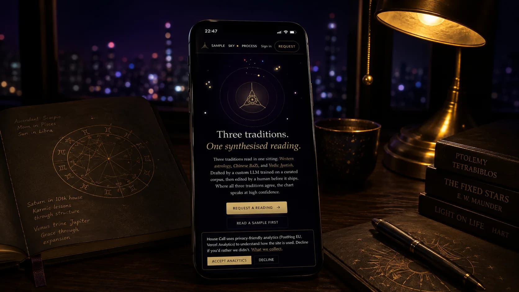A phone propped on a dark wooden desk at night, House Call's landing page glowing on screen: the gold orbital glyph inside its rings above the headline Three traditions. One synthesised reading. in serif and gold italic, the three-tradition explainer and a Request a Reading button beneath it. Beside the phone, an open notebook with a handwritten natal chart in gold ink, a ceramic cup, and a stack of astrology books (Ptolemy's Tetrabiblos, The Fixed Stars, Light on Life); a brass desk lamp glows warm at the right, blurred night-city lights in the window behind.